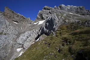 Steep mountain face with scattered snow fields and green grassy areas under a clear sky