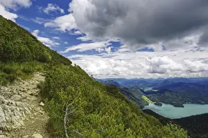 A stony hiking trail along a wooded mountain slope with sweeping views down into the valley to Lake Walchensee