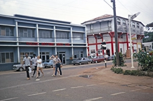 Street scene with people walking past the Follys Betting 1963 Ltd. building, Mandeville, Jamaica, 1970, Central America