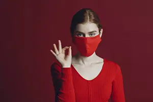 Studio portrait ok sign of young woman in protective medical face mask isolated on red background