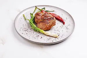 Succulent grilled meat sits on a textured plate surrounded by fresh scallions and bright red chili peppers