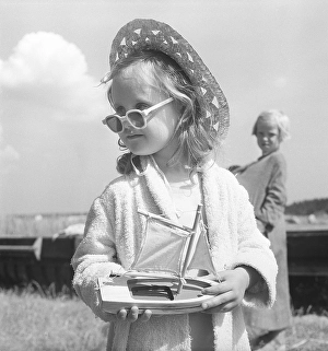 Summer of the 1950s. A young girl is playing on the beach. She is wearing a sun hat and sunglasses. In her hands a charming wooden sailing boat toy. Sweden 1953. Kristoffersson BL110-3
