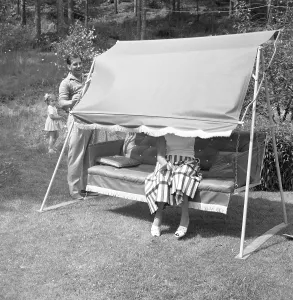 Summer in the 1950s.A woman is sitting in a hammock but her husband plays with her