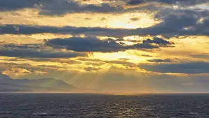 The sun sends its rays through the clouds over the sea near a mountain