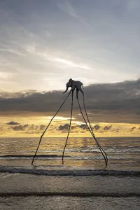 Surreal elephant sculpture with long legs by the ocean at sunset with gentle waves