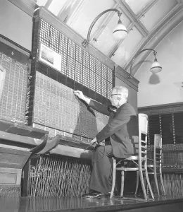 A telephone exchange. A man sits at a manual switchboard. All incoming and outgoing calls must be connected and he does this by connecting the cord and plug to the correct connection