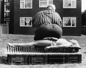 Thick man sitting on a woman lying on nail board