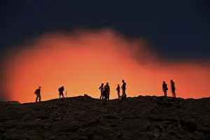 Tourists at the crater rim of the volcano Erta Ale in front of a red glowing steam cloud
