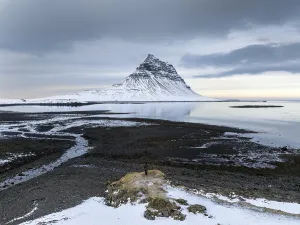 An unidentified man stands on a snow-dusted outcrop facing Kirkjufell