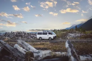 A van stands in a rural area between piles of wood and mountains in the background
