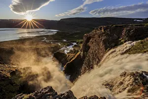 Waterfall backlit by midnight sun in front of mountains and coast