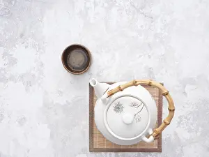 White teapot with bamboo handle and small wooden teacup on bamboo mat