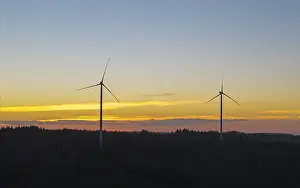 Two wind turbines in front of a glowing sunset sky over a wooded landscape