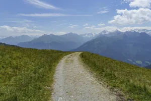 A winding mountain path leads through a green meadow with a mountain panorama in the background