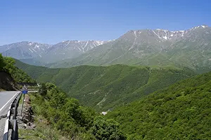 A winding mountain road snakes through green hills with mountains in the background