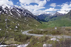 A winding road runs through a green valley between snow-capped mountains under a blue sky with white clouds