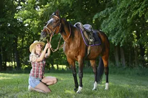 A woman (cowgirl) with a straw hat kneels beside a polish arabian horse grazing in a lush