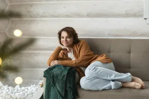A woman in a cozy brown cardigan and blue pants lounges on a gray sofa