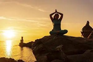 A woman doing meditation and yoga exercises at sunset next to a lighthouse in the sea