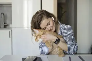 A woman embraces a ginger cat while sitting at a table in a cozy kitchen
