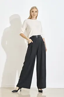 A woman in grey pants and a white top poses in a simple indoor space
