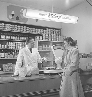 Woman in a grocery store in the 1940s A young woman stands at the counter where a merchant holds out a jar of rhubarb