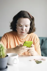 Woman at home, wearing facial beauty mask while browsing on her smartphone