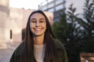 A woman with long brown hair and glasses smiles warmly outdoors She's wearing a green coat and beige sweater