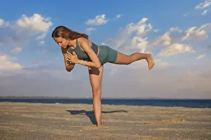 A woman performs Virabhadrasana Warrior 3 pose on the beach during sunset
