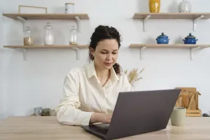 A woman sits at a table, focused on her laptop