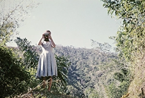 Woman taking photographs with a camera in a forested area of Trinidad, around 1962