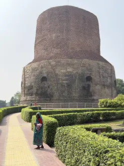 A woman in traditional dress walks past a large old brick tower surrounded by manicured hedges and a cobbled path