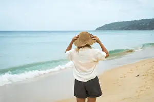 A woman, wearing a sun hat, stands on a sandy beach