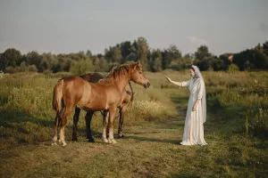 A woman in white interacts peacefully with two horses in a sunny meadow