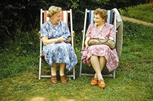 Two women friends sitting in deckchairs on grass lawn one knitting, smiling at each other, England, UK early 1960s
