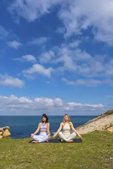Two women practicing meditation and yoga in a serene lotus pose on a yoga mat overlooking the ocean from a grassy cliff