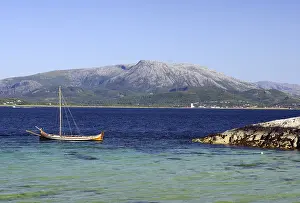 A wooden boat floats on a clear blue sea in front of a large mountain