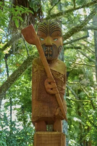 Wooden statue of a Maori with a carved face and head in a lush forest setting