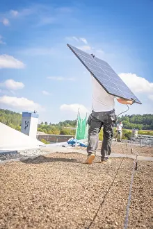A worker carries a solar panel over the roof in sunny weather and blue skies