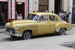 A yellow Chevrolet vintage car stands on an urban street surrounded by historic buildings