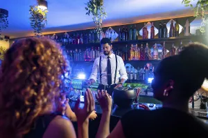 Young bartender preparing cocktails for cheering customers in a trendy bar with blue lights and hanging plants