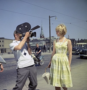 A young blonde woman is being filmed by a man with a film camera