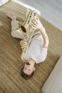 Young boy lying comfortably on a woven carpet and playing a saxophone