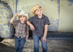 Two young boys wearing cowboy hats leaning against antique truck