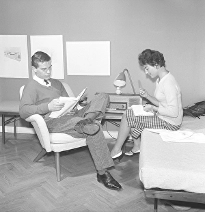 A young couple sitting together at home in the 1950s. The man sits well dressed in shirt and tie reading while the young woman sits on the bed embroidering monograms on their bedclothes and pillowcases