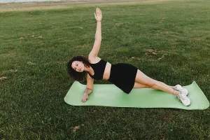 Young curly athletic girl in sportswear performs a side plank exercise on a yoga mat outdoors on the grass during dawn