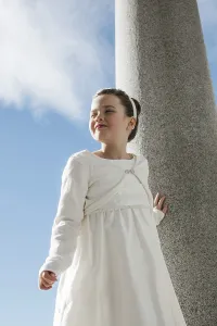 A young girl wearing a white First Communion dress stands outdoors against a stone column