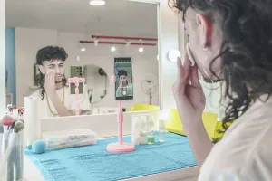 Young male beautician cleaning his face with a cotton pad while recording a makeup tutorial on his smartphone in a