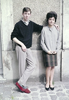 Young man casual smart clothing red slippers with his arm around shoulder of slim pretty young woman against wall of building, France c 1960