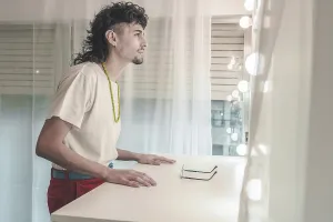 Young man with a mullet hairstyle and a colorful necklace is leaning on a dressing table with glasses on it
